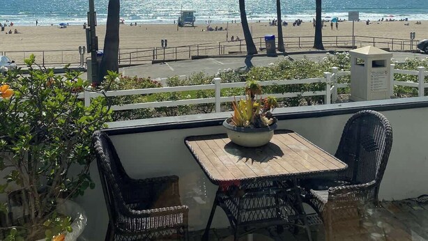 A patio with a table and chairs overlooking a beach with palm trees and the ocean. - Manhattan Beach Beachfront Charming On The Strand (Manhattan Beach)
