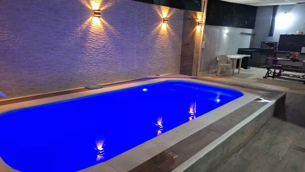 Piscina com iluminação embutida, parede de pedra e área de refeições com mesa e cadeiras.
