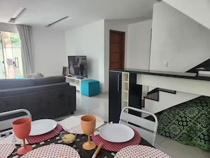 A modern living room with a sofa, a television, and a dining area with a table set for two. - HOUSE, 4 SUITES, POOL, BARBECUE, COMFORT, SPACE AND TRANQUILITY (Serra Grande Niterói)