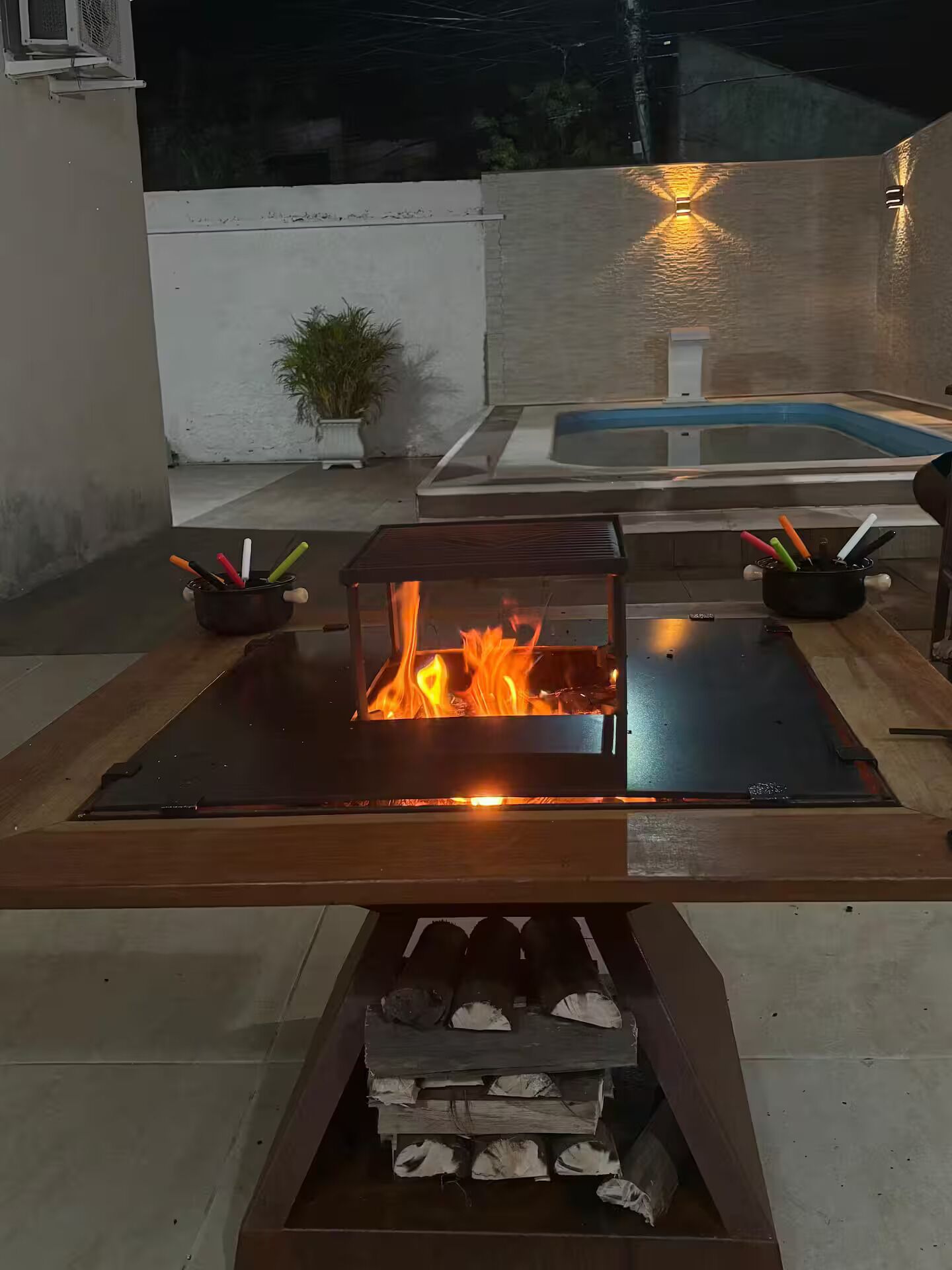 A fire feature with logs and a metal table, surrounded by a concrete patio with a potted plant and a swimming pool in the background.