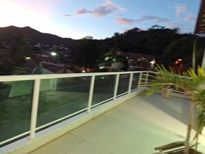A balcony with a glass railing overlooking a mountainous area with distant lights. - HOUSE, 4 SUITES, POOL, BARBECUE, COMFORT, SPACE AND TRANQUILITY (Serra Grande Niterói)
