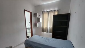 Quarto pequeno com uma cama de solteiro, estrutura de madeira, guarda-roupa, pequena estante e janela com cortinas.