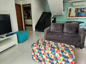 A living room with a TV, a sofa, a colorful ottoman, and a staircase leading to another level.