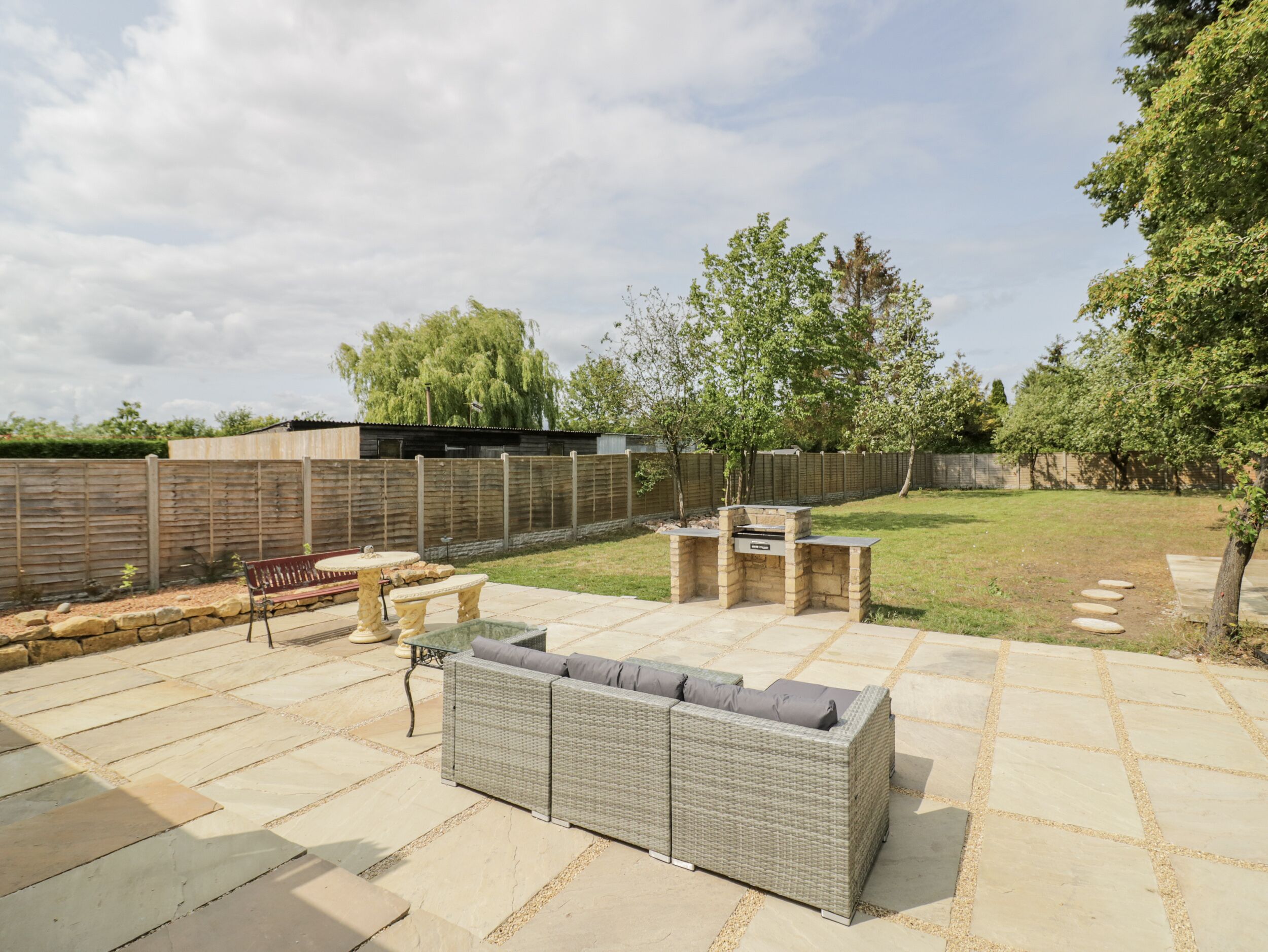 A patio area with a stone-tiled floor, a wicker sofa, a barbecue grill, and a wooden fence.