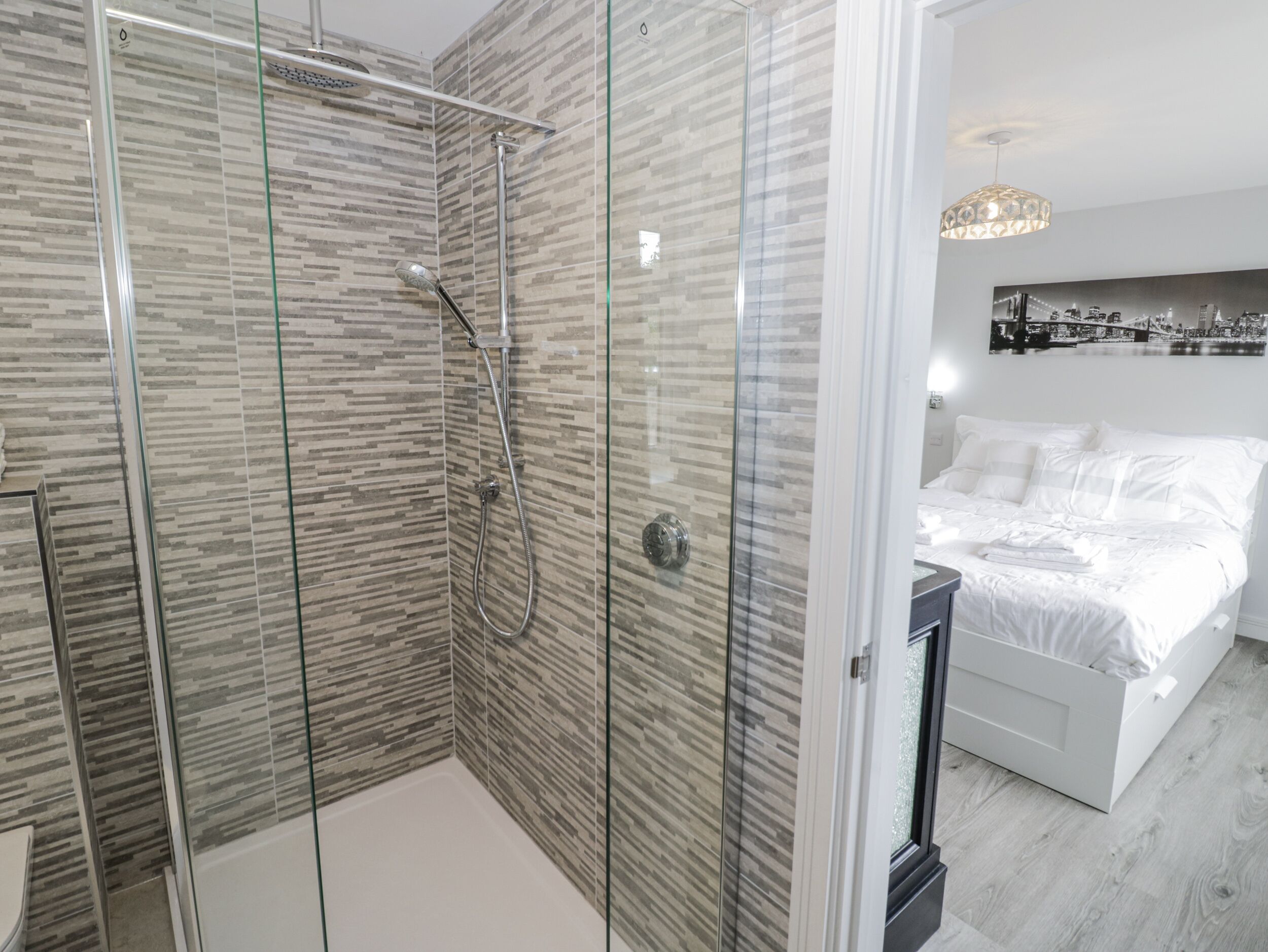 A modern bathroom with a glass-enclosed shower and a separate bedroom with a bed and a nightstand.