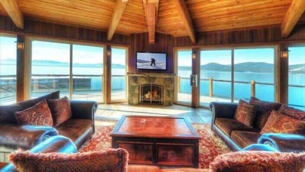 A spacious living room with a stone fireplace, large windows offering a view of the water, and wooden ceiling beams. - Bright and Cozy Lakeview Retreat #59 (Kings Beach)