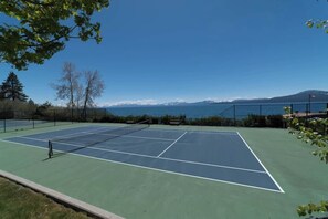 An outdoor tennis court with a net, surrounded by trees and a view of mountains and water. - Brockway Springs Lake View Unit- #30 (Kings Beach)
