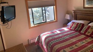 A bedroom with a bed, a window with blinds, a television mounted on the wall, and a nightstand with a lamp. - Lakefront Condo in Kings Beach-#33 (Kings Beach)