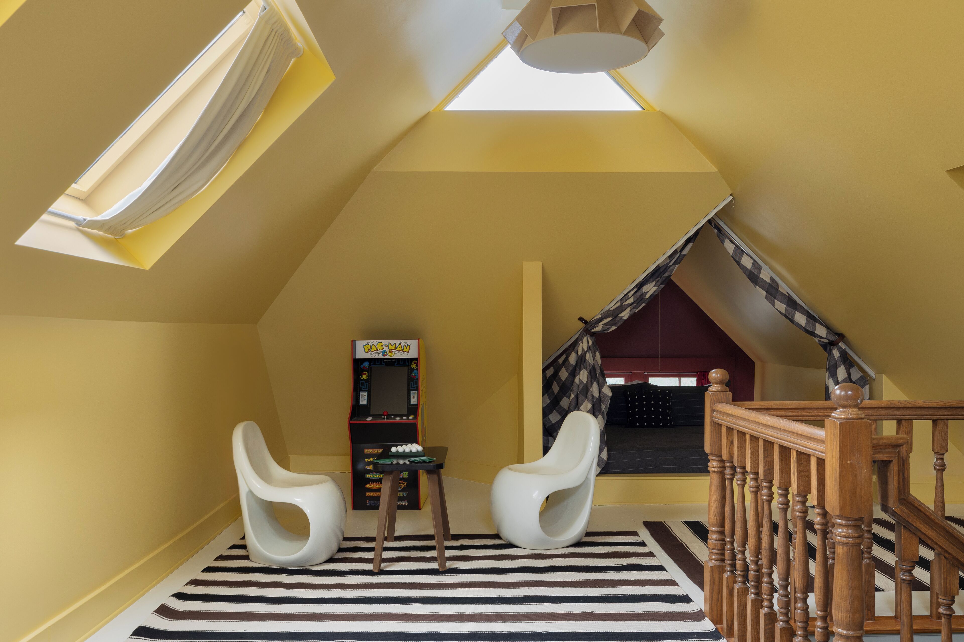 Una habitación con techo inclinado, tragaluces, una máquina arcade Pac-Man, dos sillas blancas, un tapete a rayas y una escalera de madera.