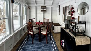 A dining area with a wooden table, chairs, and a blue rug. There are windows along one wall and a kitchen area with a coffee maker and a shelf. - Charming, lake-front, 2-bedroom cabin with AC, WiFi in superb Derby (Derby)