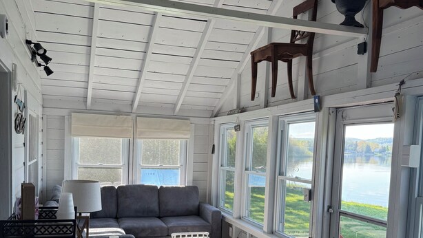 A spacious living room with a large window offering a view of a lake, a gray sofa, a coffee table, and a wooden chair. - Charming, lake-front, 2-bedroom cabin with AC, WiFi in superb Derby (Derby)