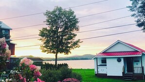 A small white cabin with a red roof, surrounded by lush greenery and flowering plants, near a body of water with a tree in the foreground. - Charming, lake-front, 2-bedroom cabin with AC, WiFi in superb Derby (Derby)