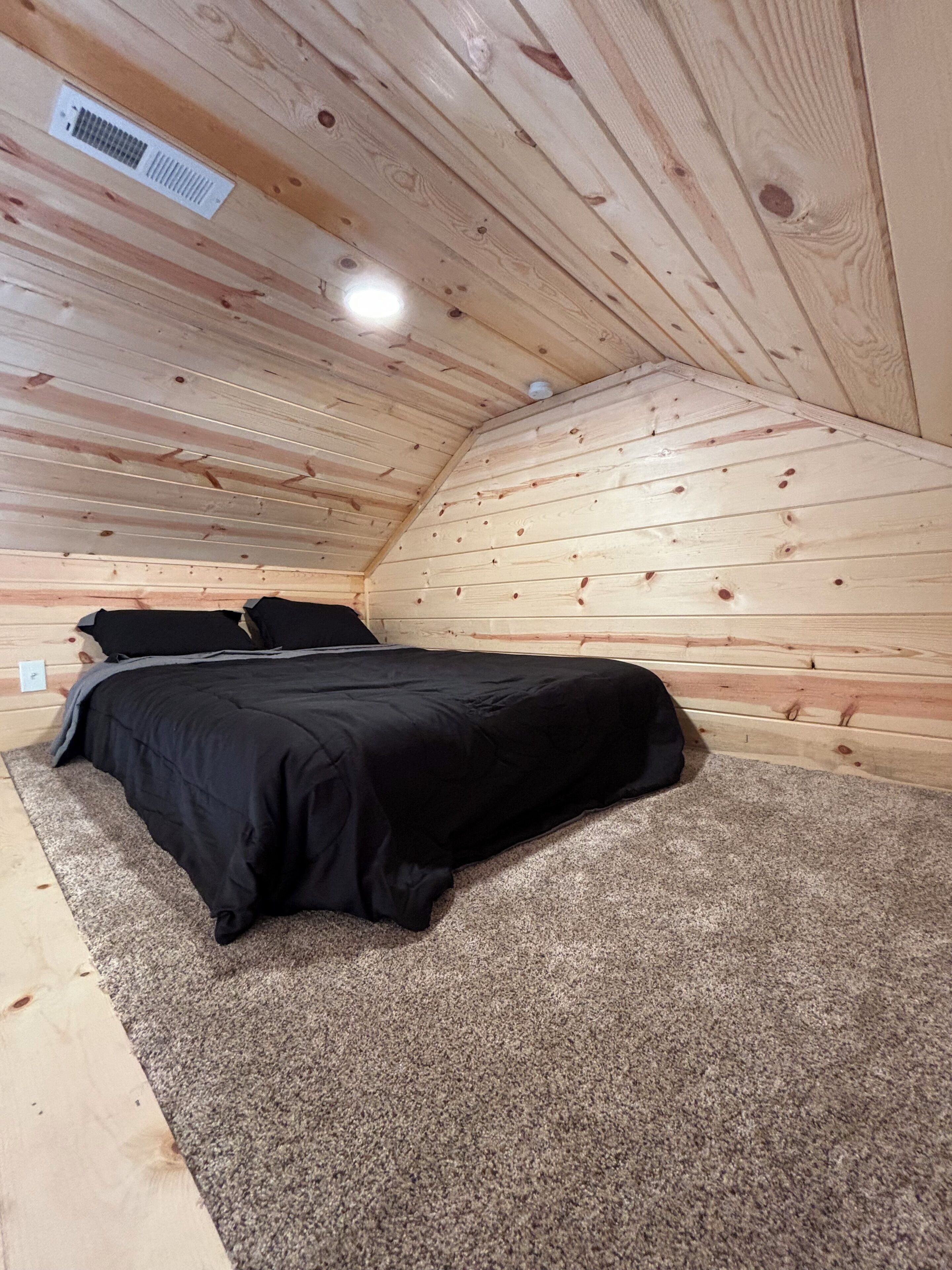 Un dormitorio con cama, alfombra, paredes de madera y un respiradero en el techo.