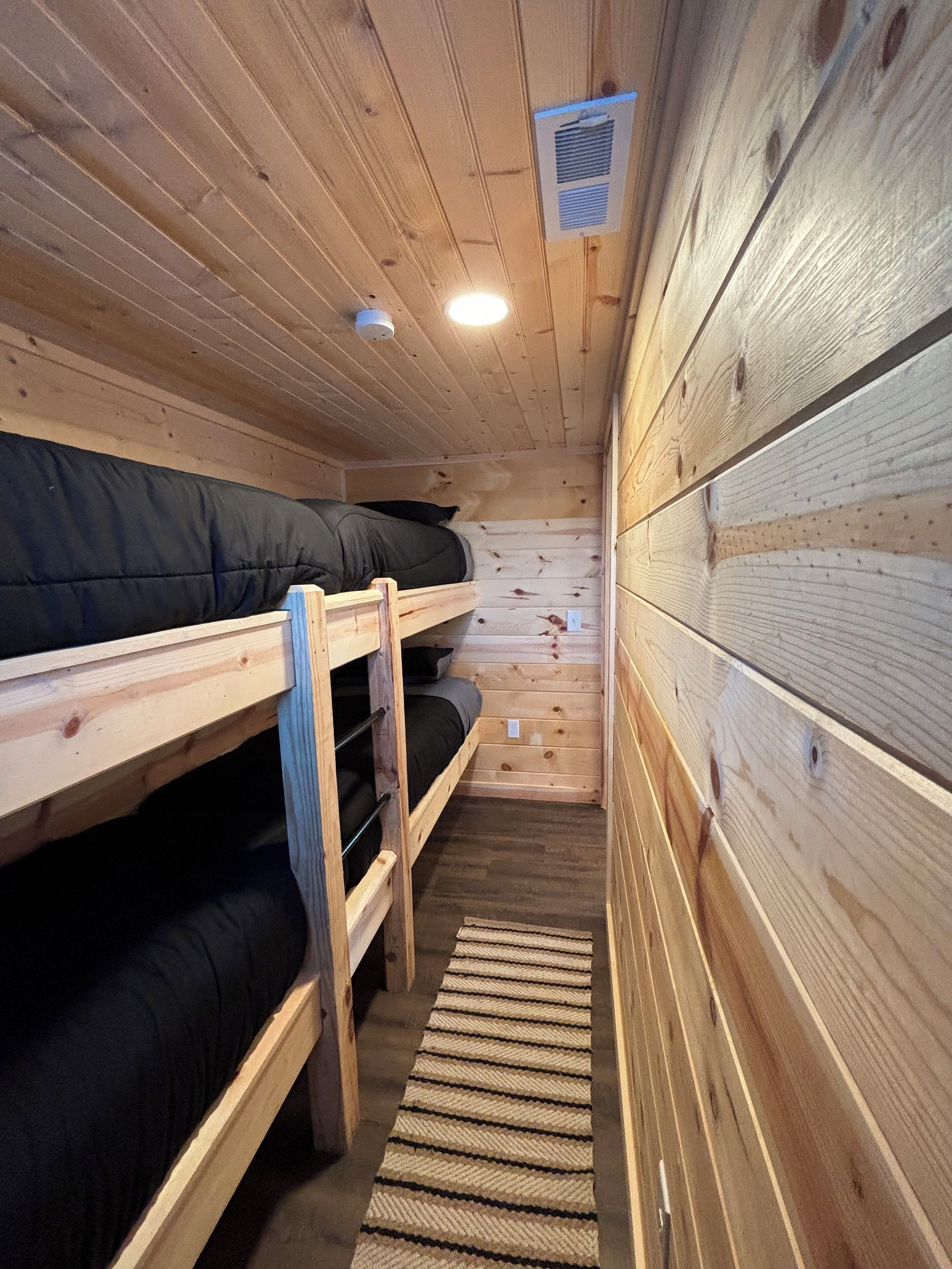 Een chalet met houten wanden en vloeren, twee stapelbedden, een klein tapijt en een ventilatie rooster.