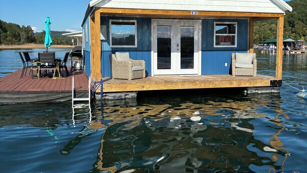 Una casa flotante con exterior azul y blanco, cubierta de madera y un letrero de 'Se vende'.