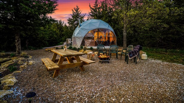 Une tente en forme de dôme géodésique, dotée d’un foyer et d’un espace salon, installée dans un cadre forestier.