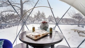 Un paysage enneigé, vu à travers une structure en forme de dôme géodésique, avec une table, des chaises et une bouteille de cidre.