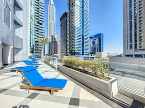 A rooftop lounge area with blue sun loungers, a tiled floor, and a view of tall buildings.