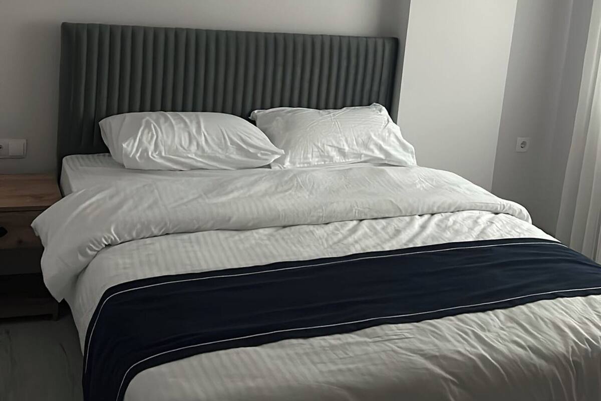A modern hotel bedroom with a large bed, a grey headboard, and a white wall.