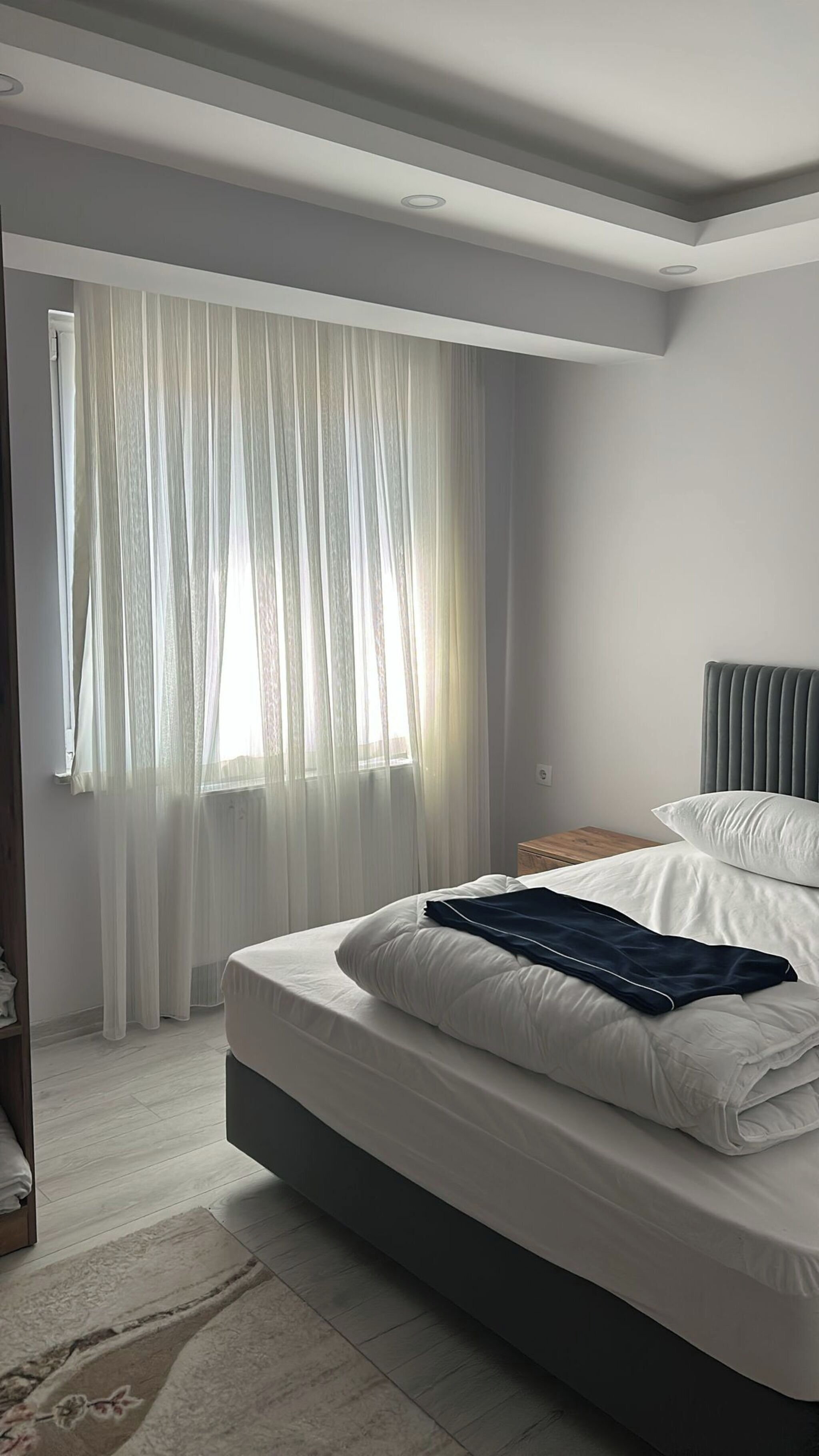 A bedroom with a bed, a window with sheer curtains, a bedside table, and a headboard.