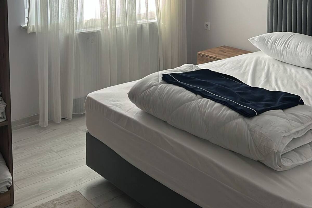 A bedroom with a bed, a window with sheer curtains, a bedside table, and a headboard.