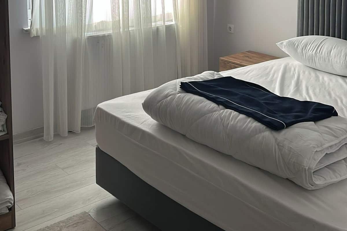 Une chambre à coucher avec un lit, une fenêtre avec des rideaux légers, une table de chevet et une tête de lit.