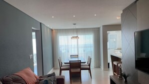 A modern living room with a sofa, a dining table with chairs, a TV, and a large window with curtains. - Apto Frente Mar Próximo ao Beto Carrero (Penha)
