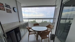 A balcony with a round table and chairs, a small kitchen area with a refrigerator, and a view of the sky and trees. - Apto Frente Mar Próximo ao Beto Carrero (Penha)