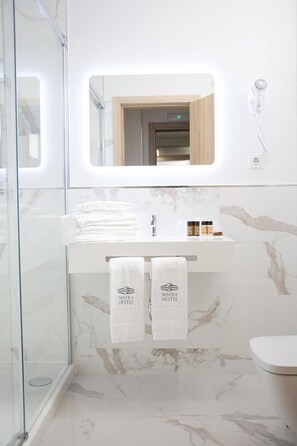 A modern bathroom with a glass shower enclosure, a marble-tiled wall, a white sink with a mirror above it, and two towels hanging on a rack. - Hotel Castelao (Mafra)