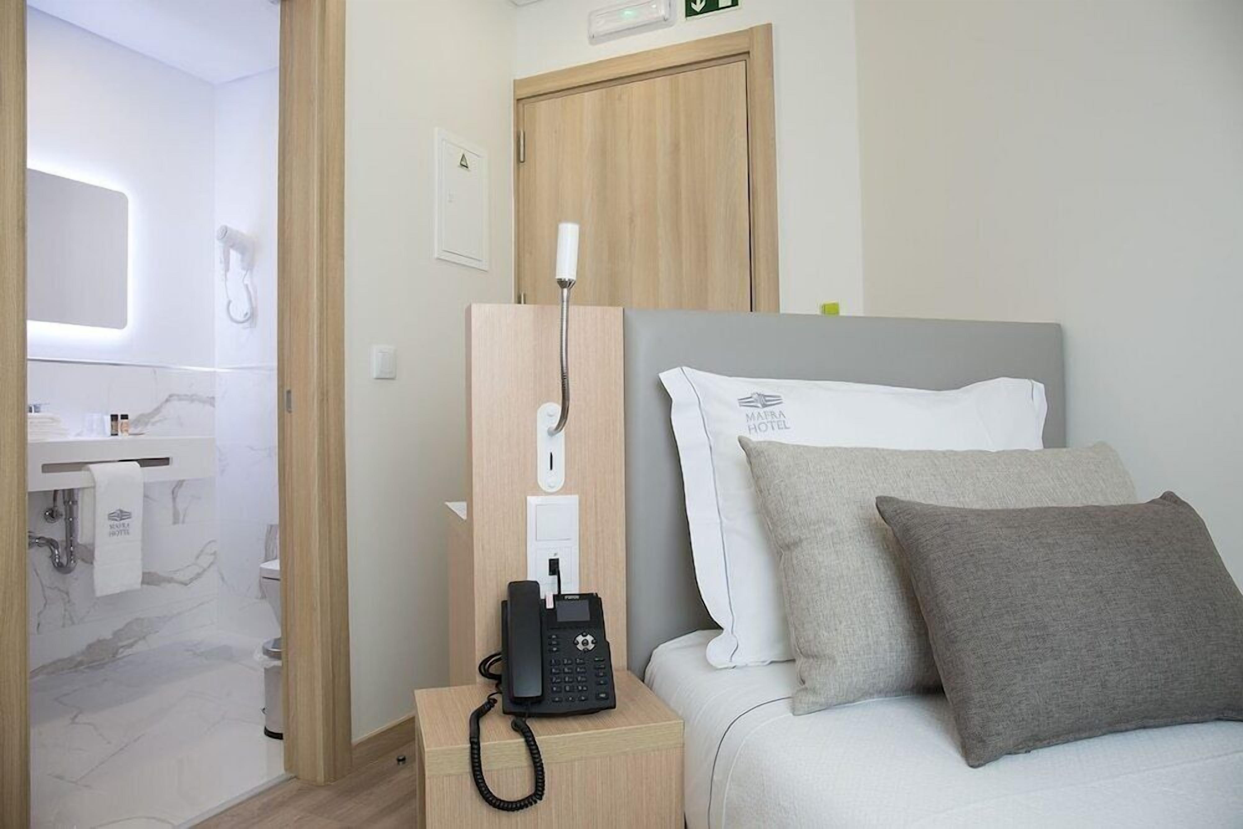 Habitación de hotel con cama, teléfono, mesita de noche, espejo, toalla y puerta.