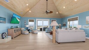 A spacious living room with a high ceiling, wooden flooring, a large white sofa, a television mounted on the wall, and a view through the glass doors.