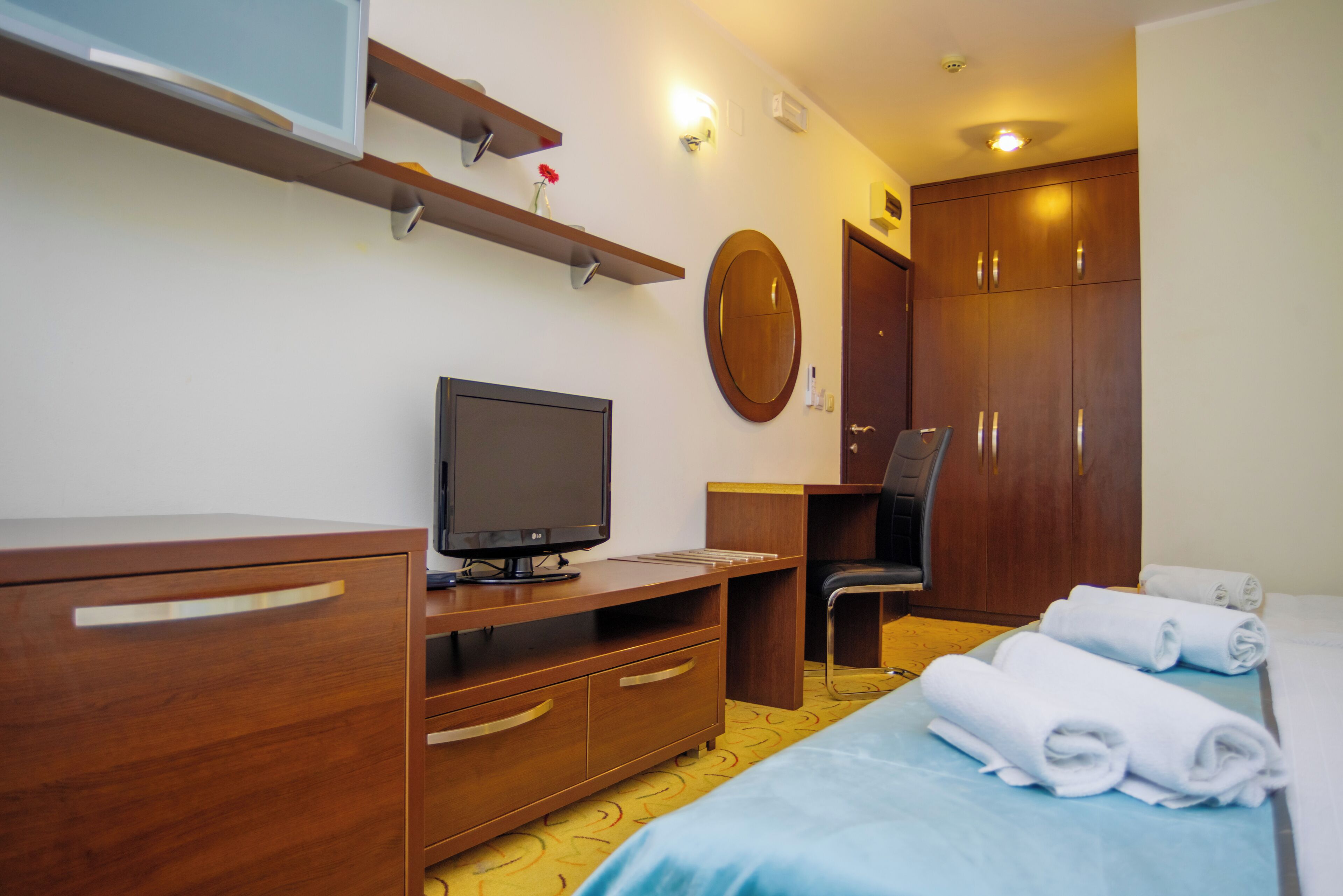 A hotel room with a bed, a desk with a computer, a TV, a wardrobe, and a mirror.