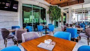 Uma área externa moderna com assentos em azul e cinza, mesas de madeira e uma grande planta em vaso.