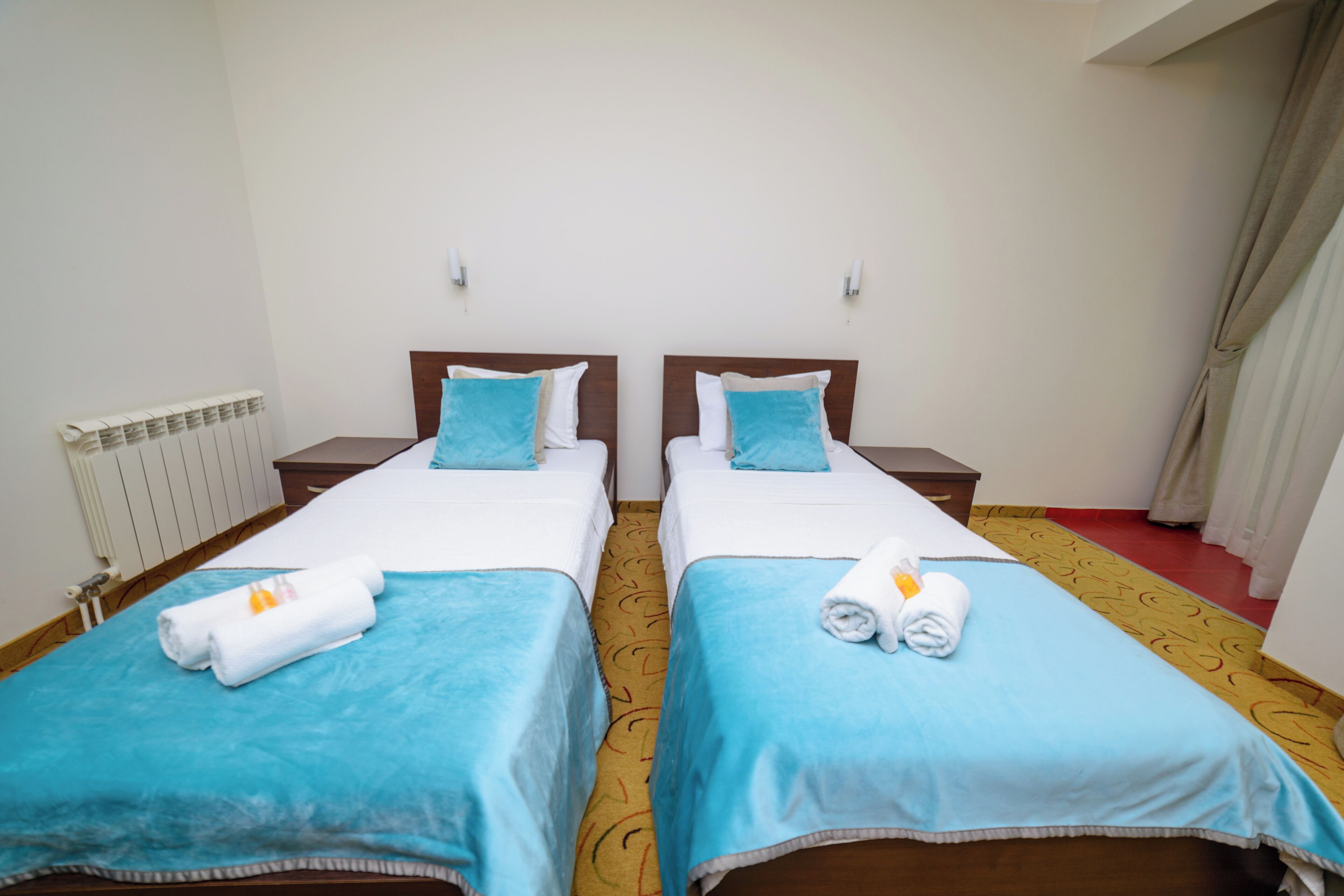 A hotel room with two single beds, each with a blue bedspread and white pillows. There is a radiator on the left wall and a window with curtains on the right.