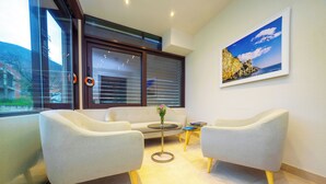 Quarto moderno com duas poltronas cinzentas, uma mesa redonda pequena com um vaso e uma janela grande com persianas.