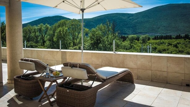 Uma varanda com móveis de vime, uma mesa com uma bebida e um grande guarda-sol.