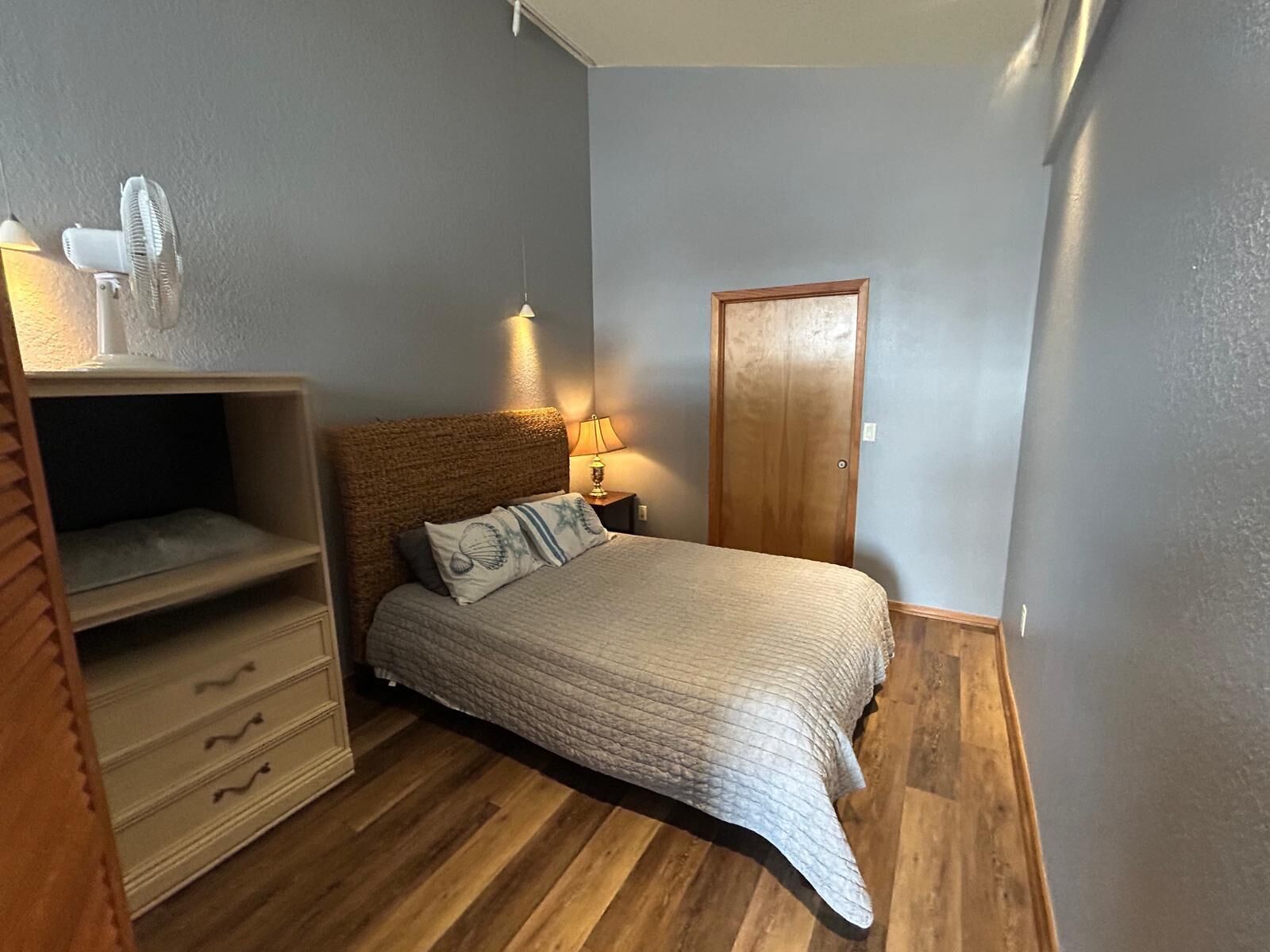Et soveværelse med seng, trægulv, et natbord med lampe, et garderobeskab og en ventilator i loftet.