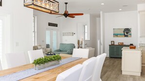 Amplia sala de estar con mesa de comedor de madera, sillas blancas, ventilador de techo y zona de cocina al fondo.