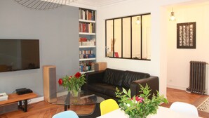 Una sala de estar con un sofá de cuero negro, una mesa de centro de vidrio con flores, una estantería, un televisor de pantalla plana y un radiador.