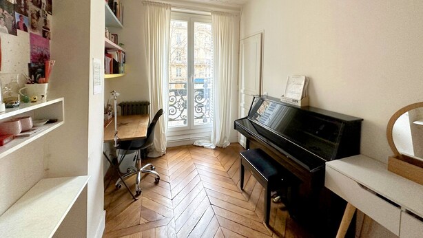 Una habitación con un piano, un escritorio y una silla, una estantería y una ventana con cortinas.