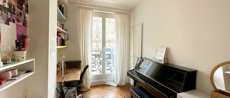 Una stanza con un pianoforte, una scrivania con una sedia, una libreria e una finestra con tende.