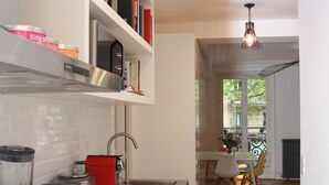 Una cocina moderna con estufa integrada, cafetera y tetera roja. Se ve un comedor con mesa y sillas a través de un puerta abierta.