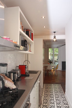 Una cucina moderna con piano cottura integrato, macchina del caffè e bollitore rosso. Si intravede un'area pranzo con tavolo e sedie attraverso una porta aperta.