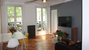 Un comedor con una mesa, sillas y una mesa baja de vidrio. Hay un radiador, una ventana con vistas y un ventilador de techo.