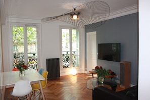 Zona pranzo con tavolo, sedie e tavolino in vetro. Si nota un termosifone, una finestra con vista e una ventola a soffitto.
