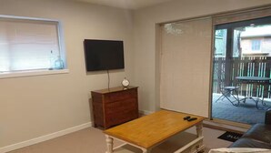 Un salon avec une table basse en bois, un téléviseur fixé au mur, une commode en bois et une porte-fenêtre coulissante donnant sur une terrasse extérieure.