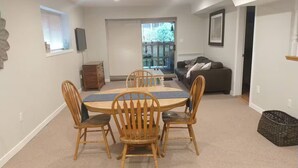 Un coin repas comprenant une table ronde et quatre chaises, un canapé, un téléviseur et une fenêtre avec des stores.
