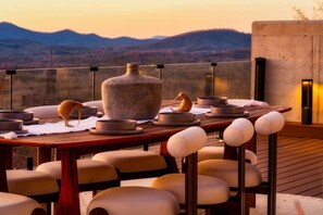 A dining area with a table set for two, featuring a large stone vase and several bowls, overlooking a mountainous landscape.