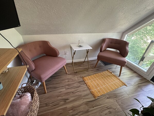 Habitació acollidora amb un sofà rosa, un butaca a joc, una taula petita blanca i un gran finestral amb vista a vegetació.