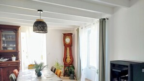 Un coin repas comprenant une table en bois, des chaises, un luminaire suspendu, une horloge comtoise et un téléviseur.